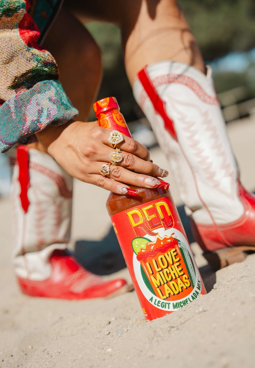 Person holding a bottle of I Love Micheladas Red Mix on a sandy surface with red Hija de Tu Madre boots